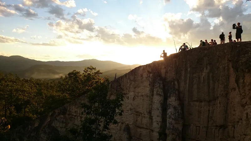 Golden sunset over Pai Canyon, with visitors on the sandstone ridge and forested hills beyond.