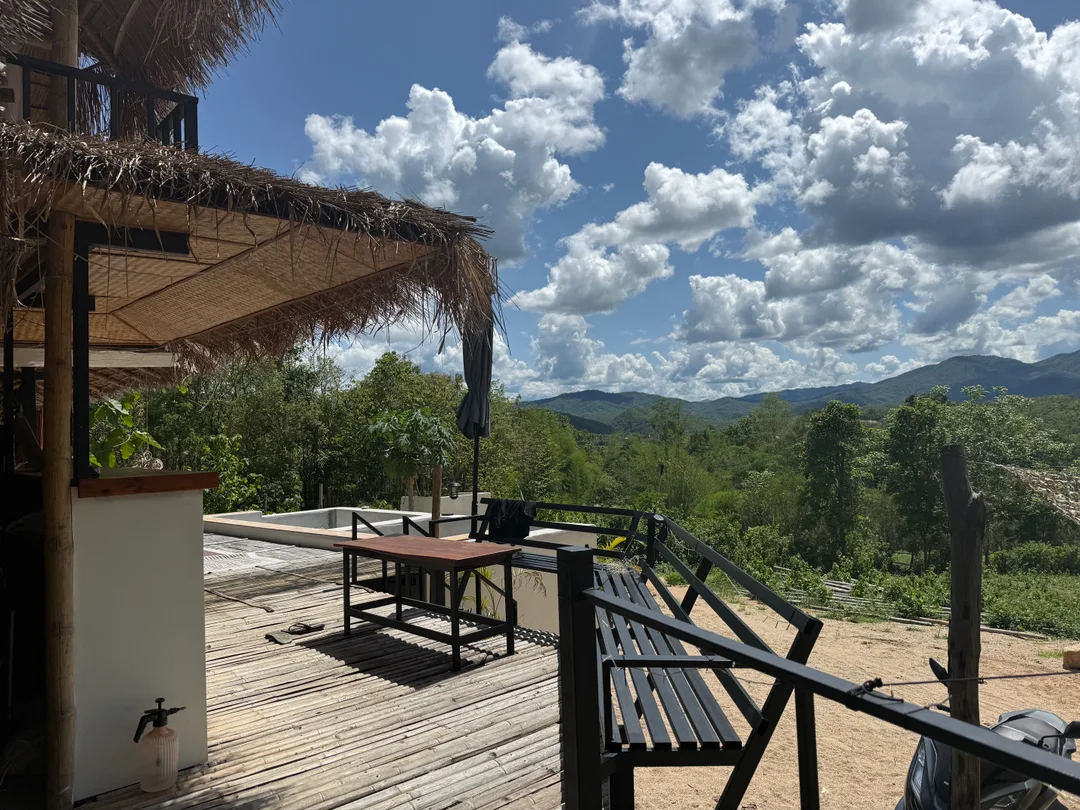 Oasis tiki bar’s thatched terrace, wooden table and seating overlooking green hills beneath a blue, cloud-filled sky.